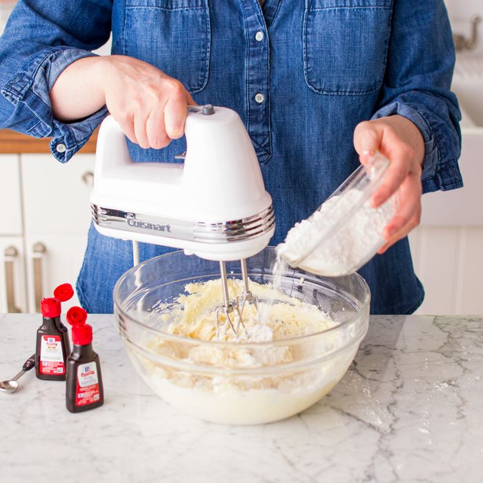 Adding Flour into a Large Glass Bowl While Mixing Ingredients with Hand Mixer on Marble Kitchen Top