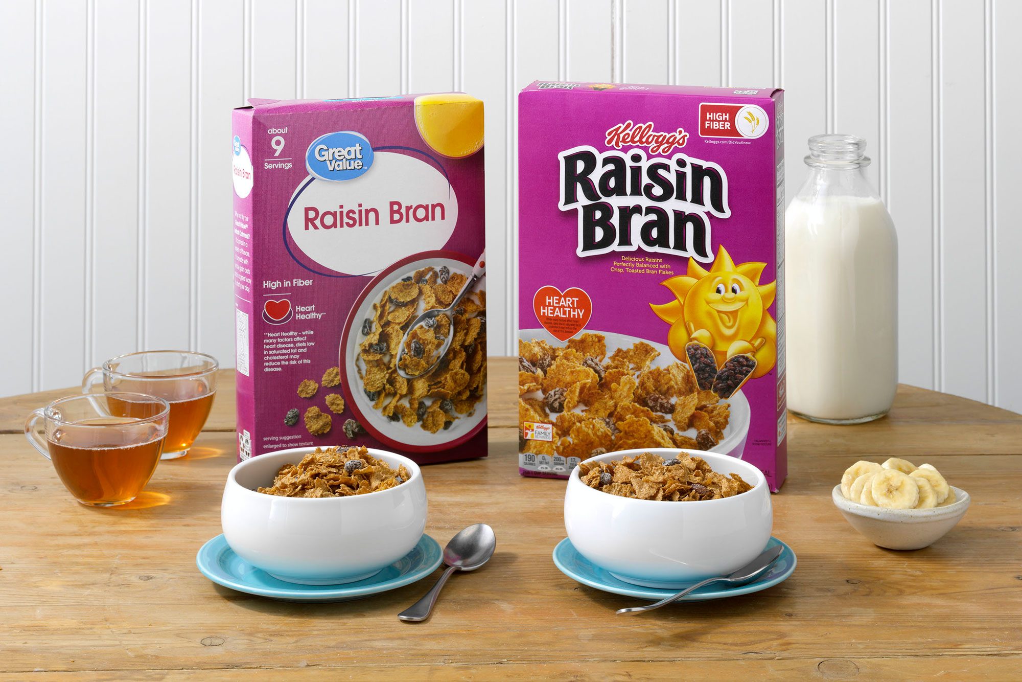 Kellogg's Raisin Bran and Walmart's Great Value Raisin Bran on a table with bananas, milk and tea