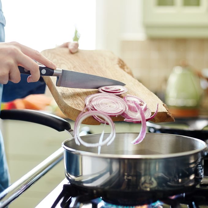 Midsection image of woman pouring chopped onions in saucepan. Utensil is placed on gas stove. Close-up of female holding cutting board and kitchen knife. She is preparing food in domestic kitchen.