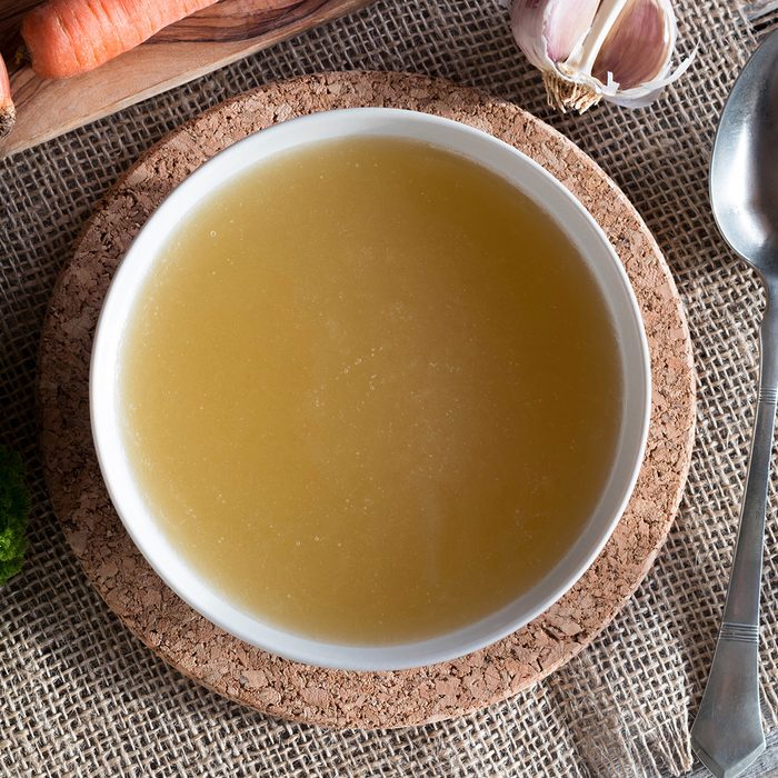 Chicken bone broth served in a soup bowl, with onions, carrots, parsley and garlic in the background