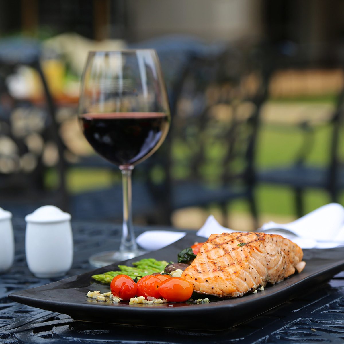 Grilled Salmon with Cherry Tomatoes and a Glass of Red Wine atop Ornamented Black Metal Table with Blurred Chairs in the Background