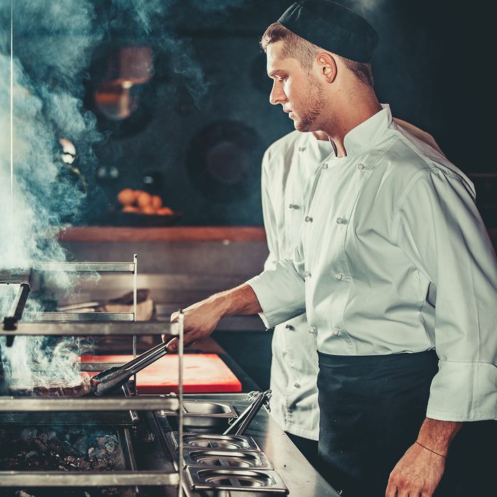 Young white chef in black apron and hat standing near the brazier whith coals.