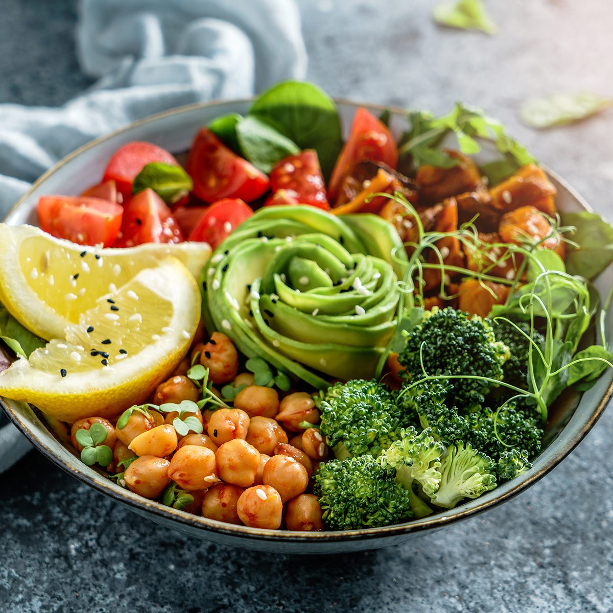 Buddha bowl salad with baked sweet potatoes, chickpeas, broccoli, tomatoes, greens, avocado, pea sprouts on light blue background with napkin.
