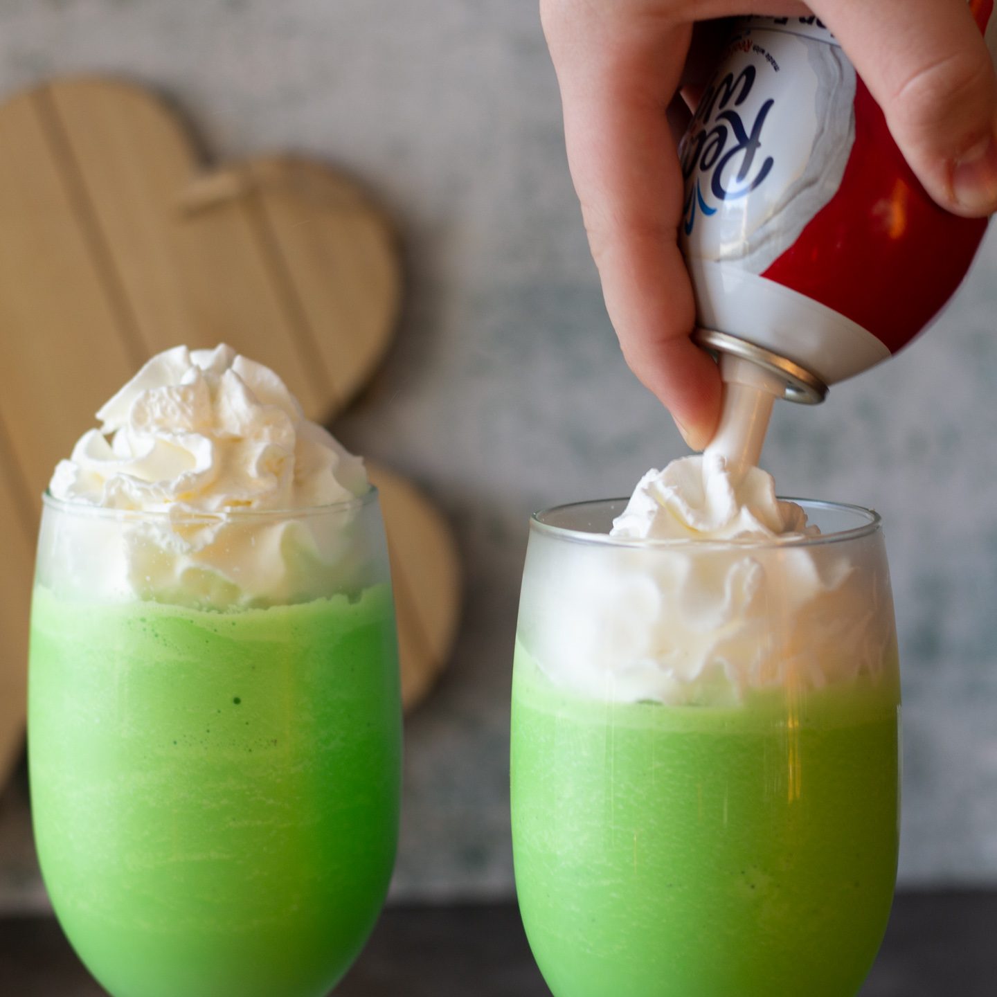 Two glasses with green milkshakes and whipped cream being added to tops.