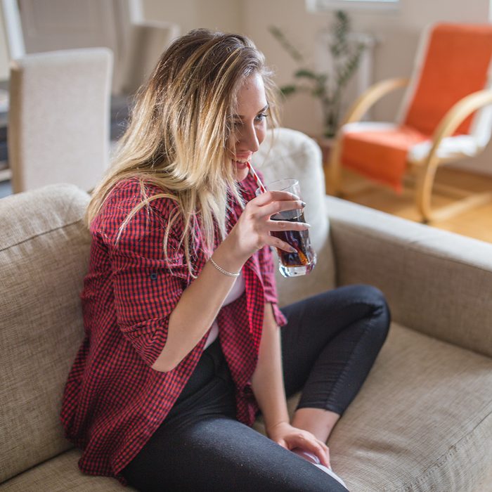 A beautiful blonde girl sitting on a sofa and drinking juice using straw. She is smiling and happy, dressed casually. She is 20 years old.