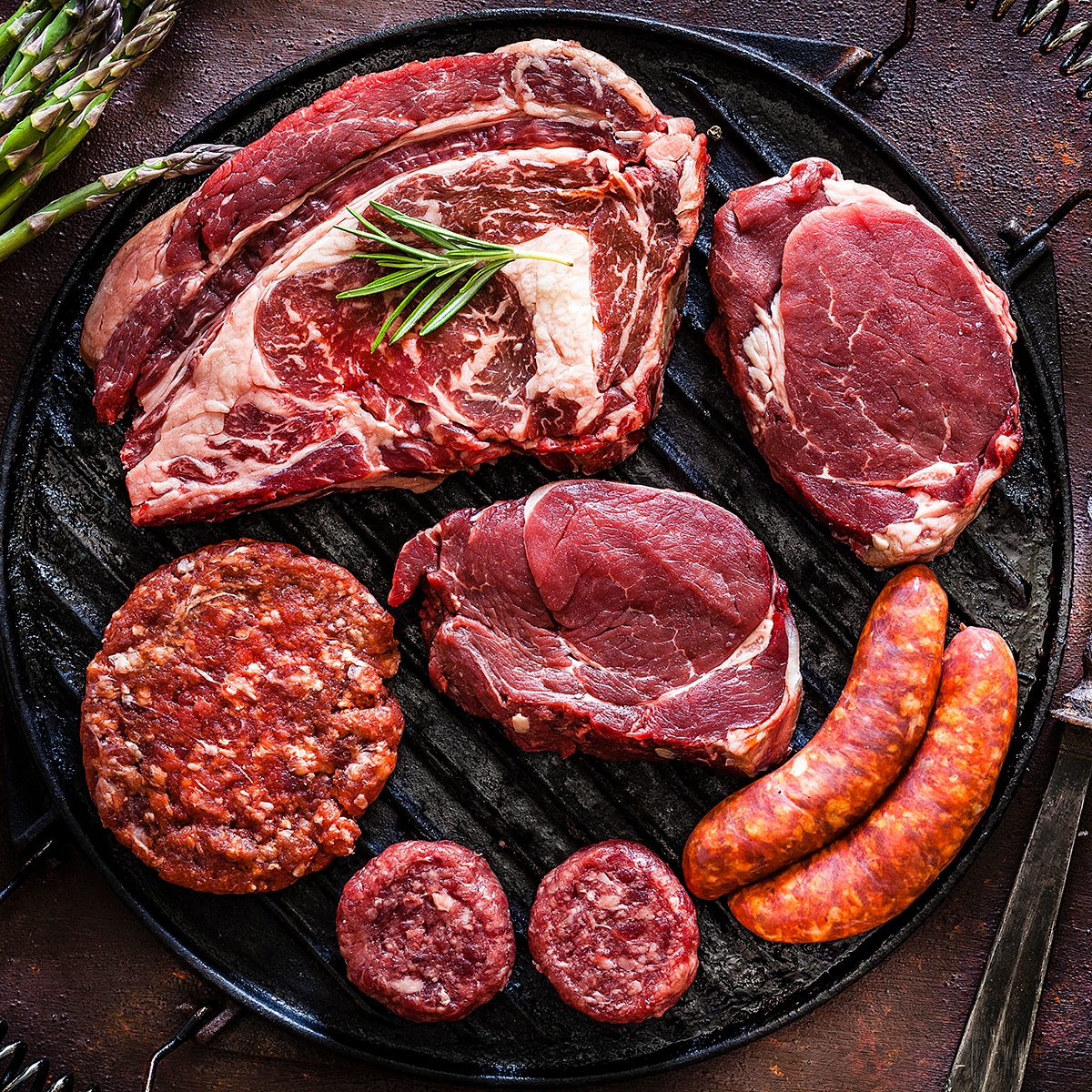 Various cuts of raw meat shot from above on rustic kitchen table. The cuts are on a cast iron grill and includes Angus steak, tenderloin, sausages and hamburger meat. The grill is surrounded by herbs, vegetables and spices for cooking meat like peppercorns, salt, garlic, tomatoes, asparagus and rosemary. A vintage fork is included in the composition. Predominant colors are red and brown. Low key DSRL studio photo taken with Canon EOS 5D Mk II and Canon EF 100mm f/2.8L Macro IS USM.