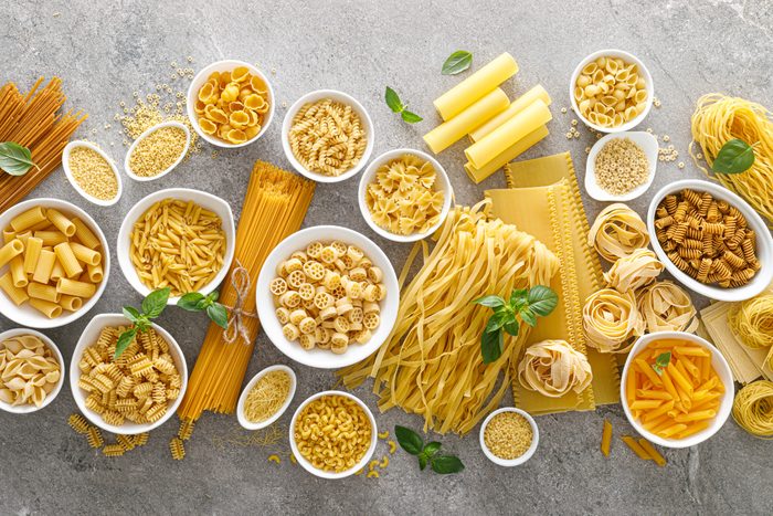 Various kinds of uncooked pasta and noodles, some in white bowls, on stone background, top view