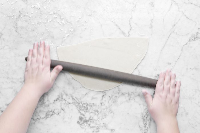 A person's hands roll out dough with a rolling pin on a marble surface