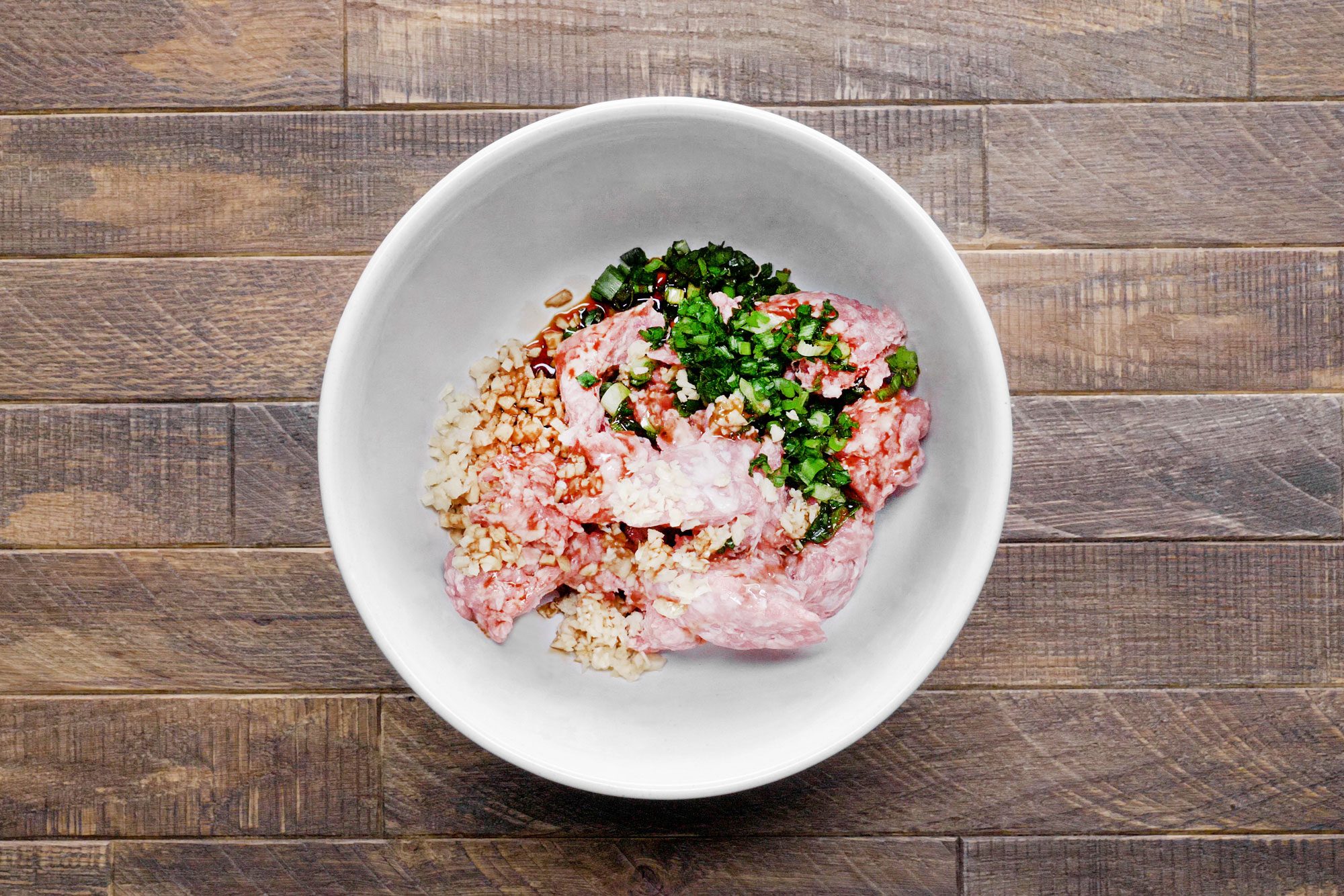 ground pork, minced green onions, garlic and fresh ginger with sesame oil and soy sauce in a large white bowl on wooden surface