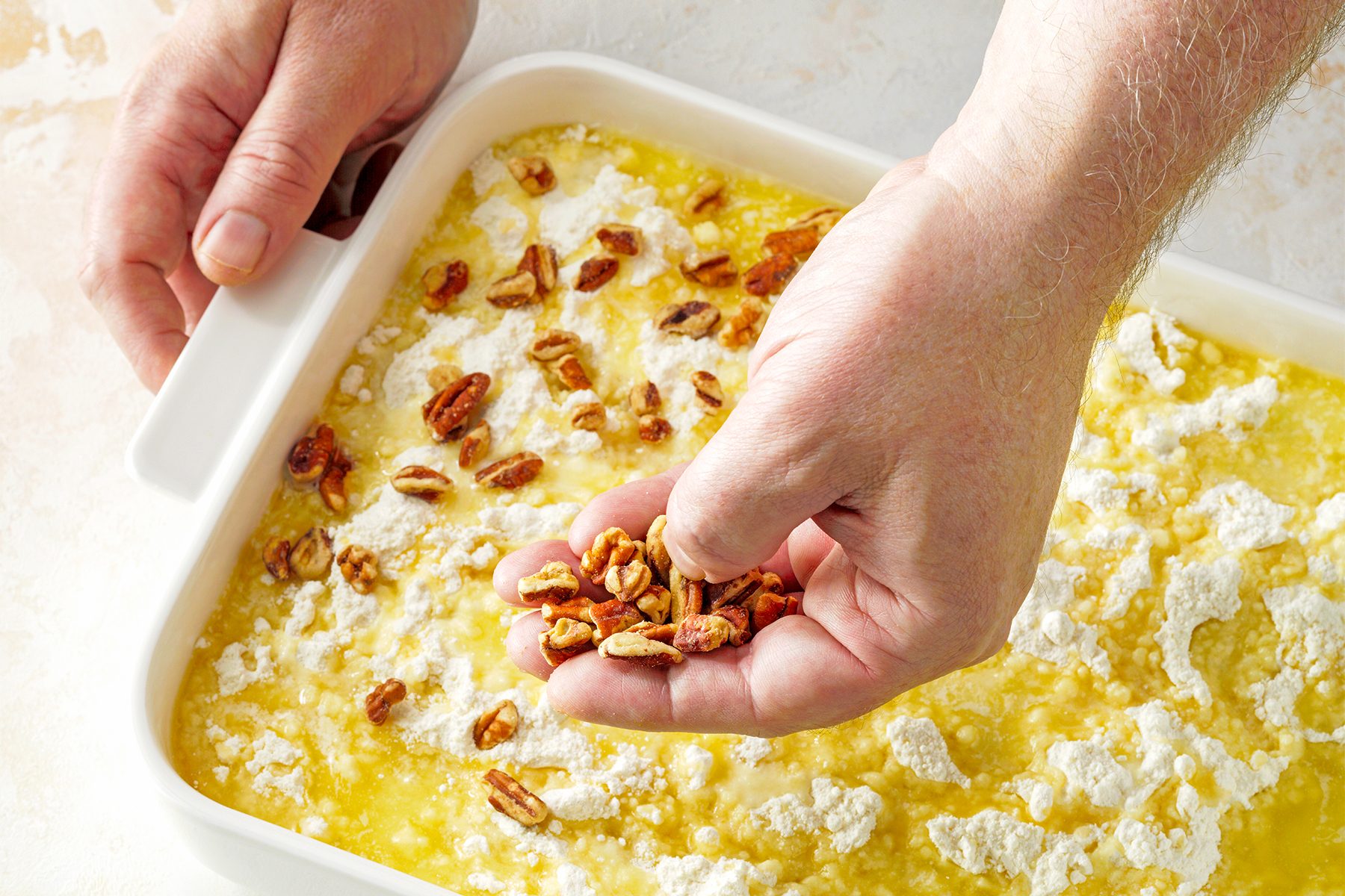 A person's hands are sprinkling pecans over a partially prepared dish in a white rectangular baking pan. 