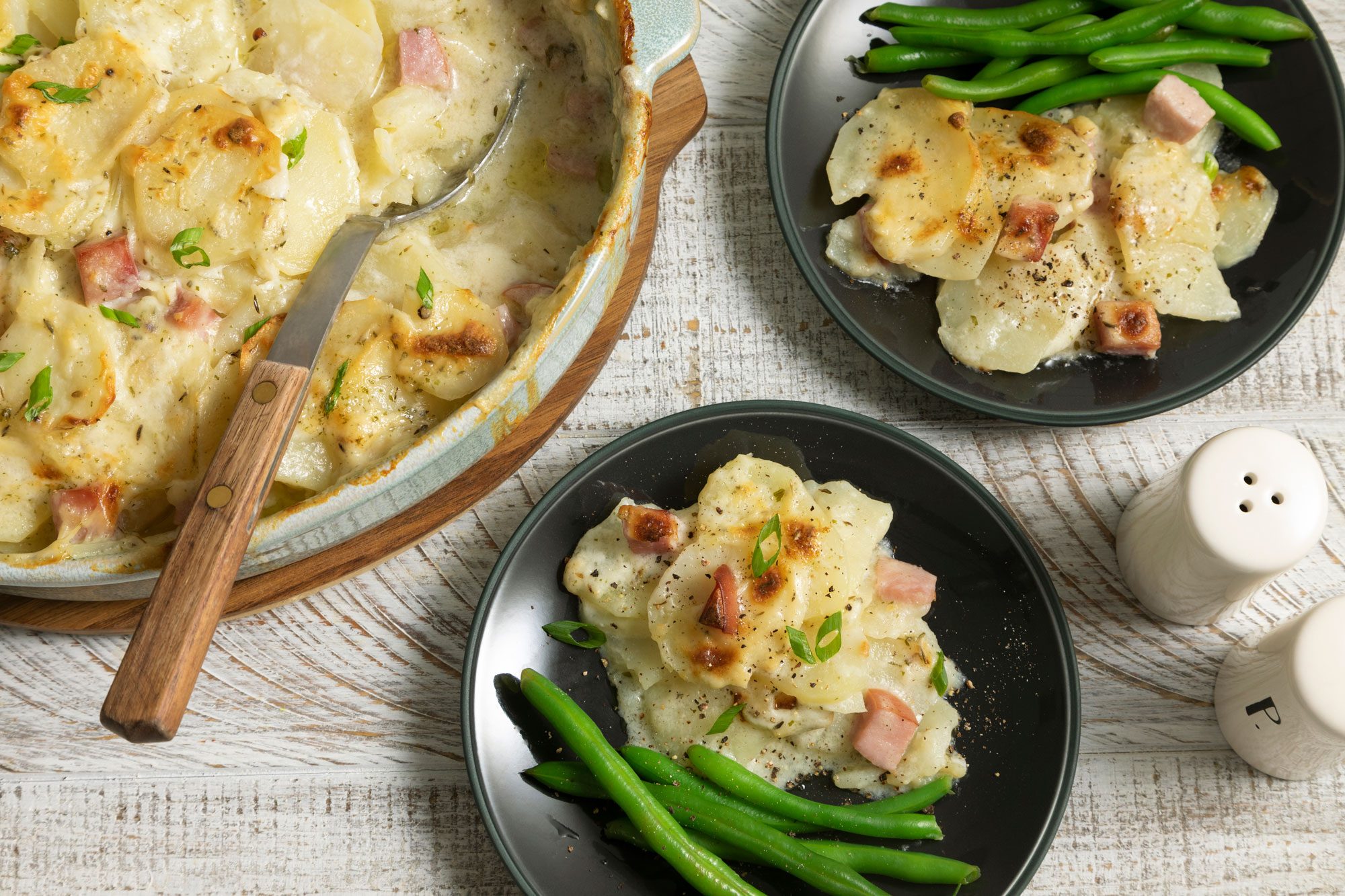 Scalloped Potatoes And Ham Served With Green Beans on a Small Black Plate on a White Painted Wooden Surface