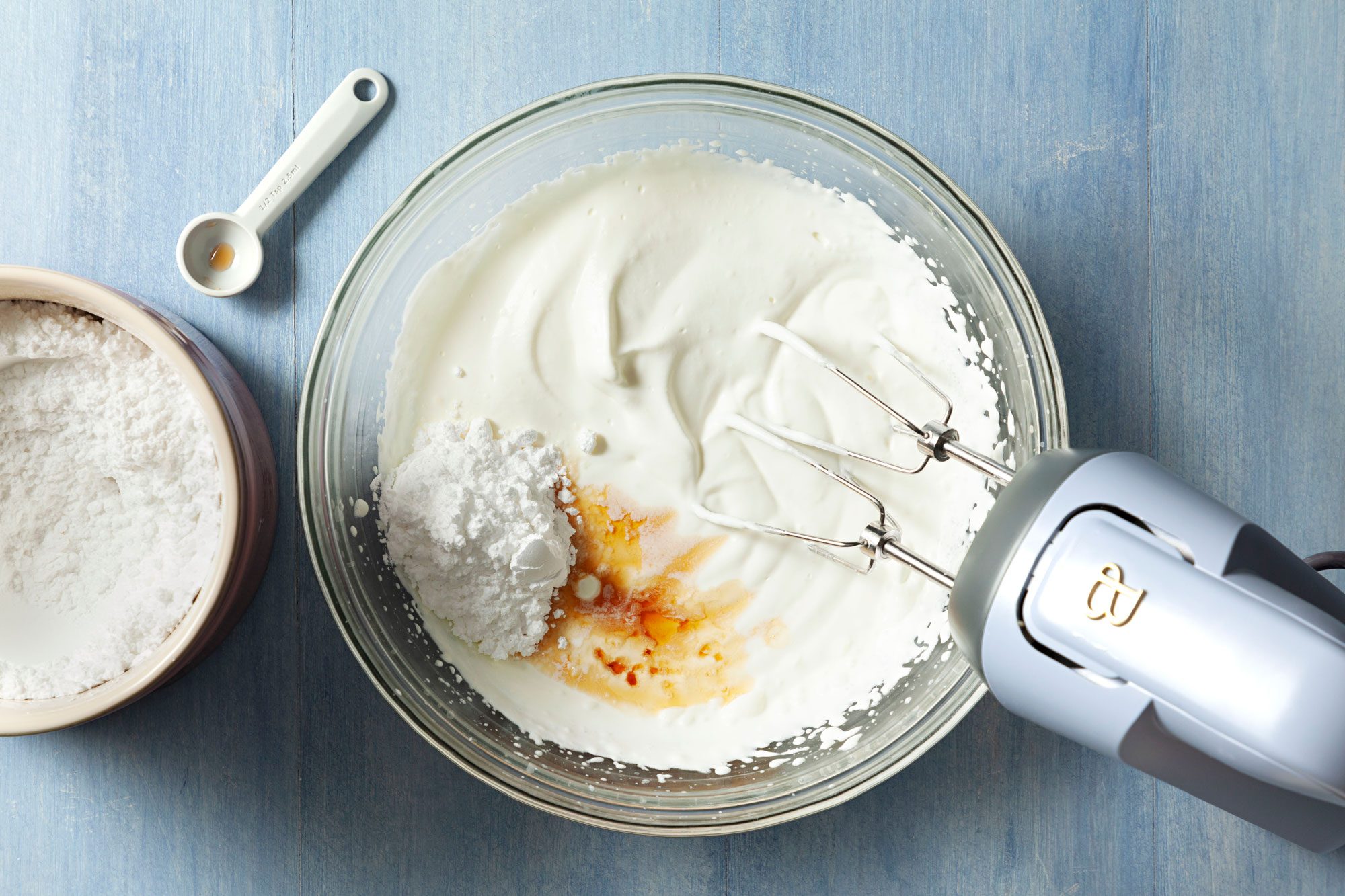 beating cream, sugar and vanilla In a large glass bowl using a hand mixer on a textured light blue surface