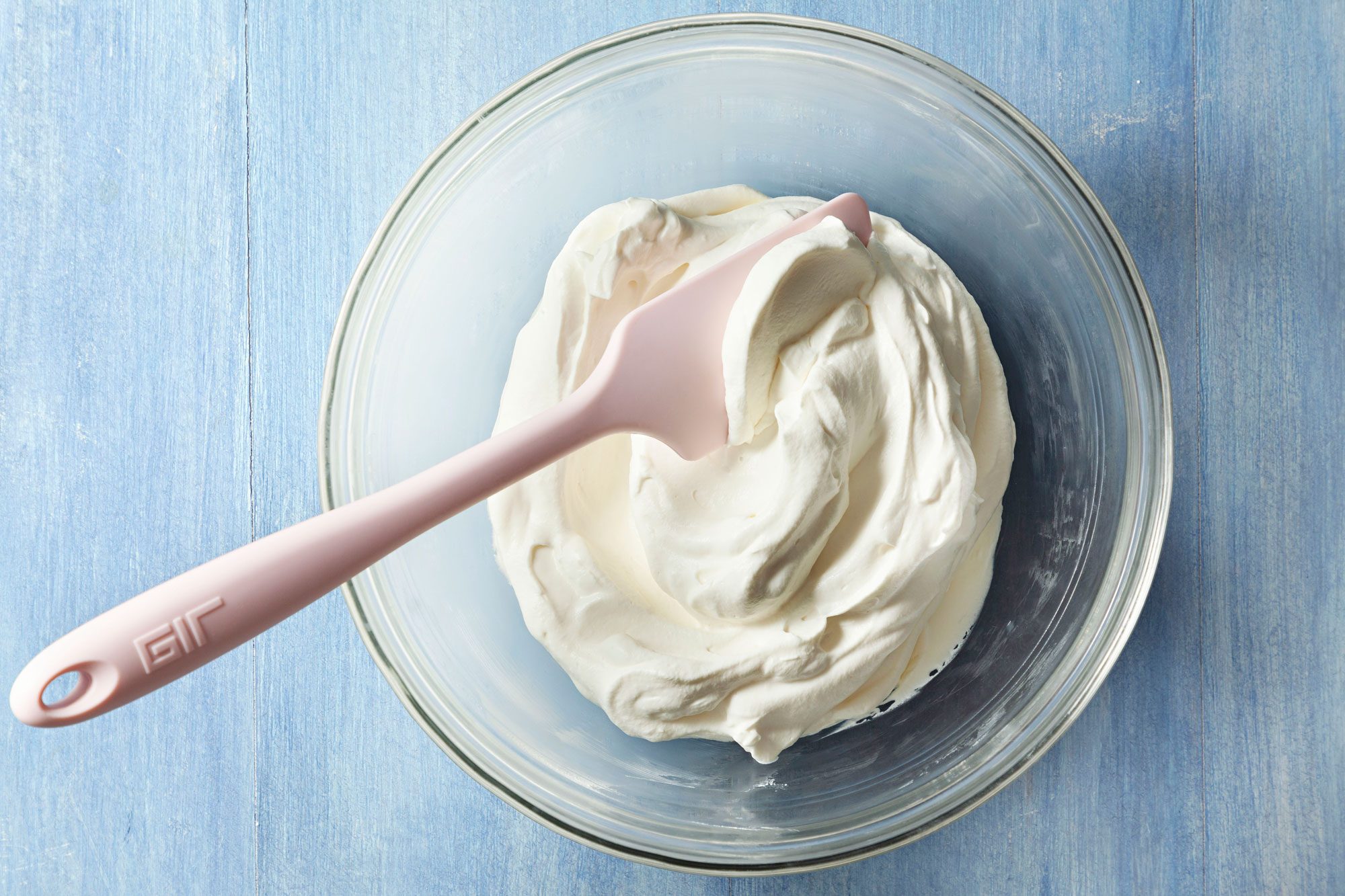 Sweetened Whipped Cream with a spatula in a large glass bowl on a textured light blue surface