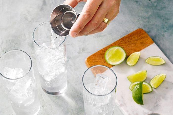 Overhead shot of fill highball or collins glasses with ice; pouring gin; lime wedges over chopping board; marble background;
