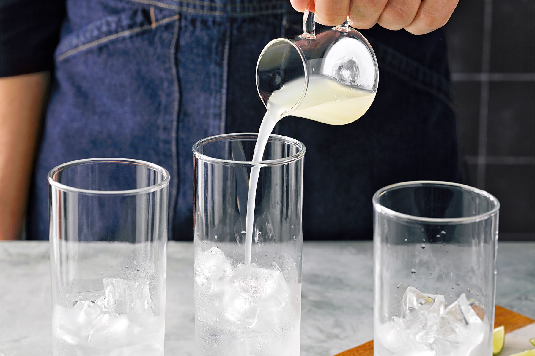 Table view shot of pouring lime juice into glasses;; lime wedges over chopping board; marble background;