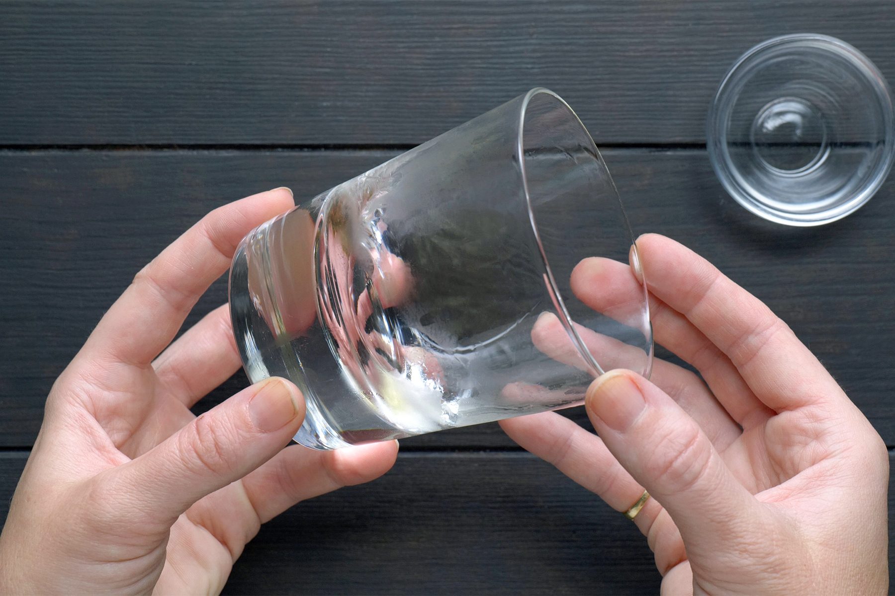 Two hands holding a clear, empty glass tilted sideways over a dark wooden surface. 