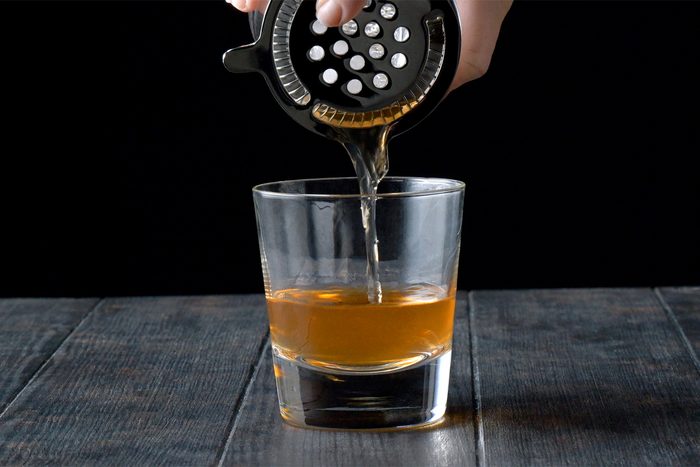 A hand holding a cocktail shaker with a strainer pours a light brown liquid into a clear glass filled halfway, set on a dark wooden table against a black background.