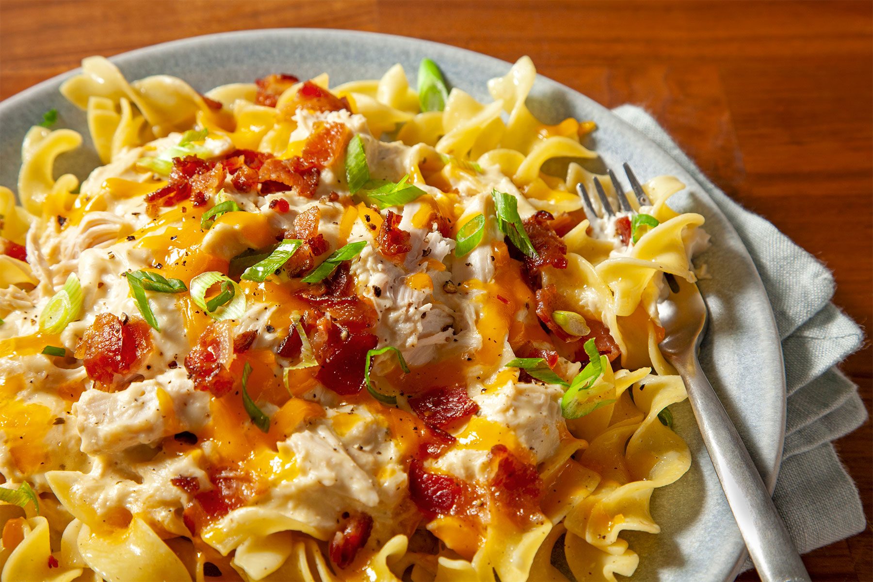 Close shot of Slow-Cooker Ranch Chicken; served on two ceramic plates; over rice or noddles; topping with green onion; fork; napkin; wooden background;