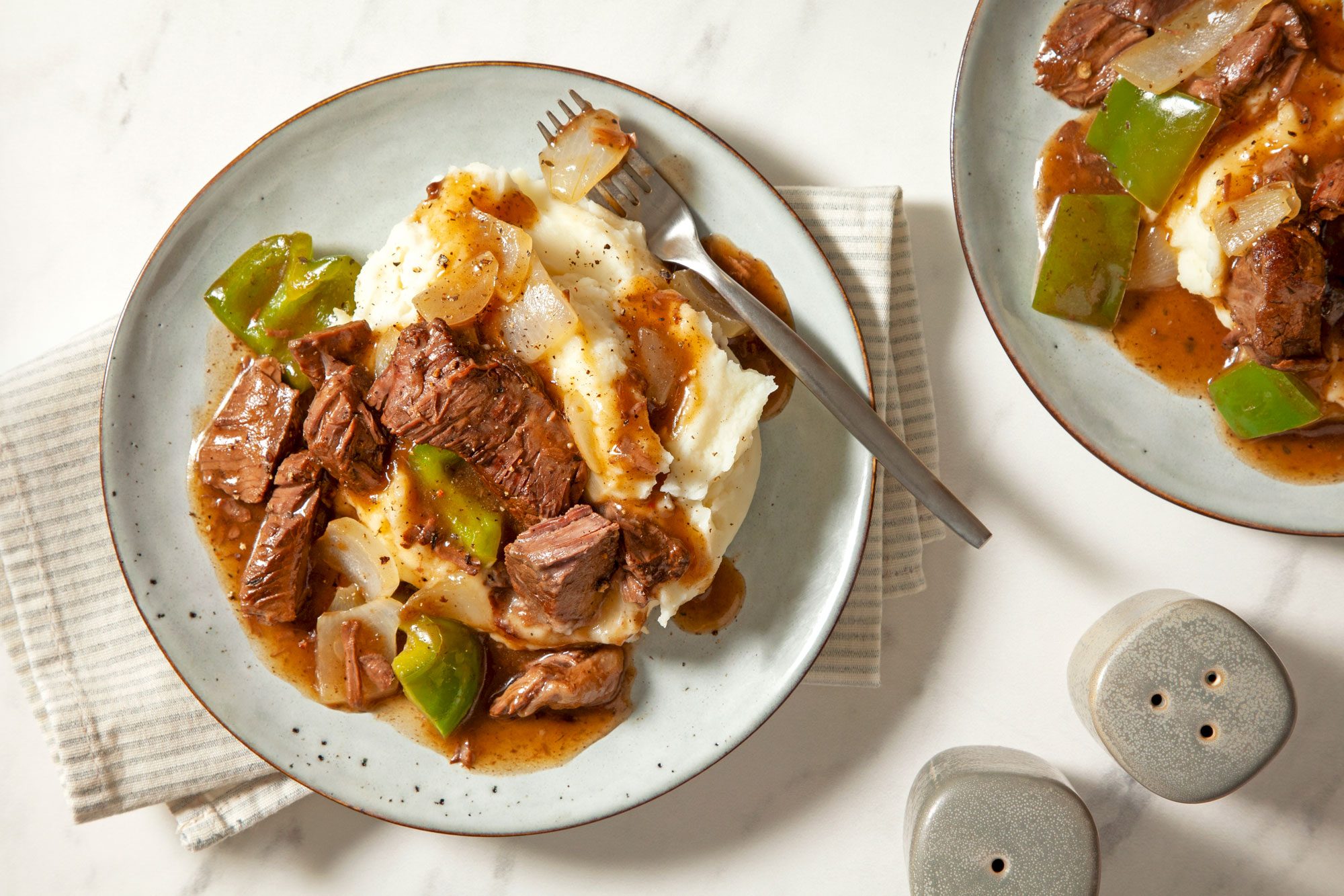 Overhead shot of Slow-Cooked Sirloin; served on two ceramic plates; salt and pepper shaker; fork; napkin; cream marble background;