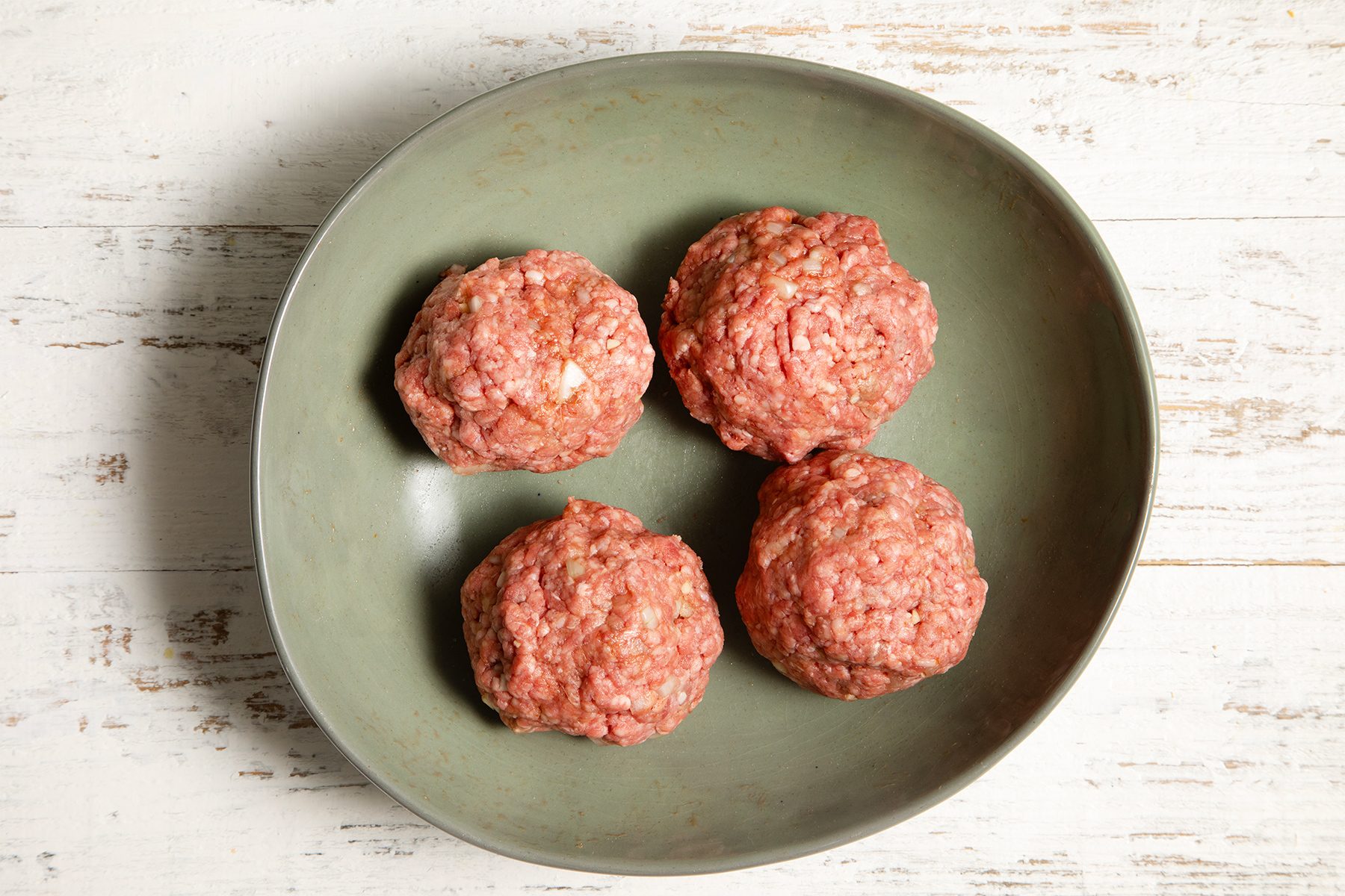 A green bowl contains four raw, seasoned meatballs placed on a white wooden surface. 