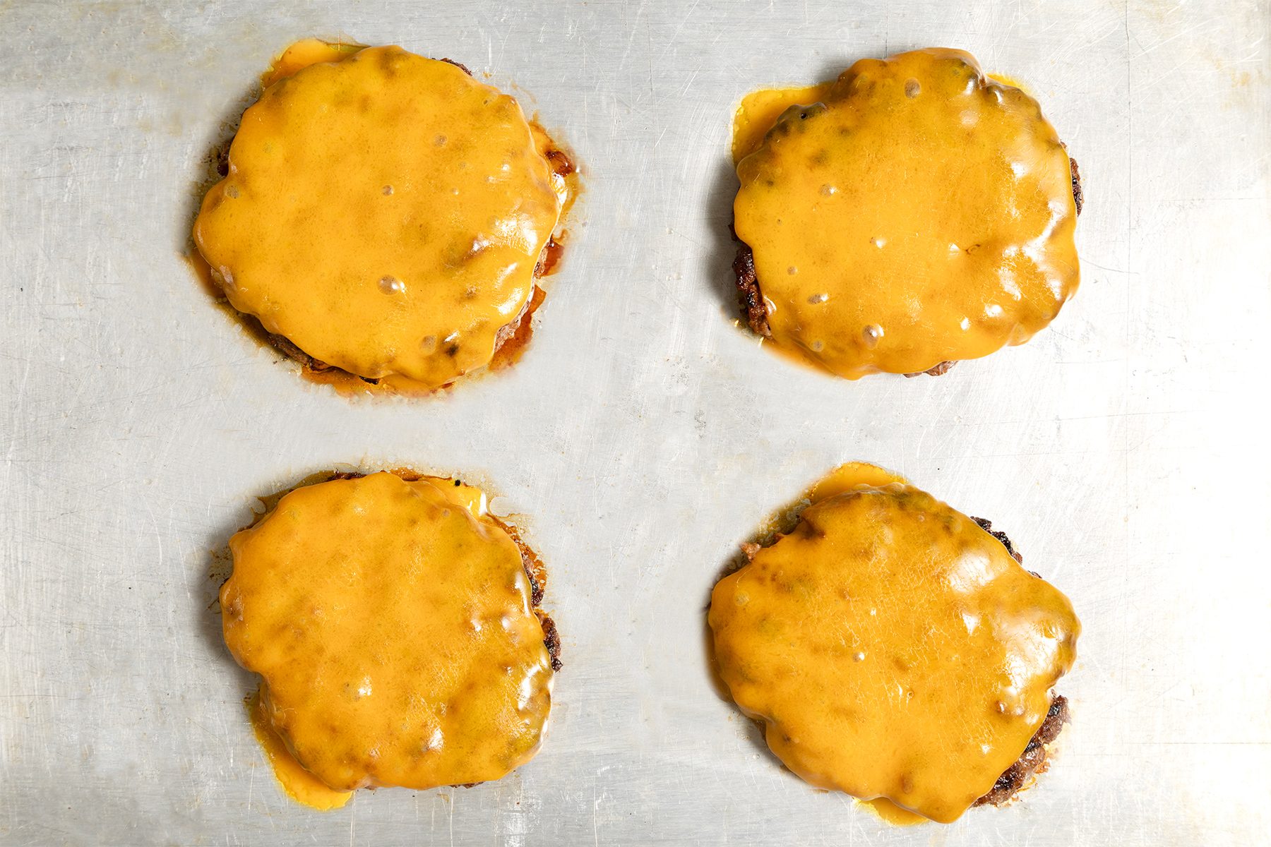 Four cheeseburgers on a metal baking sheet. Each burger is topped with a melted slice of cheese, covering the patties. 