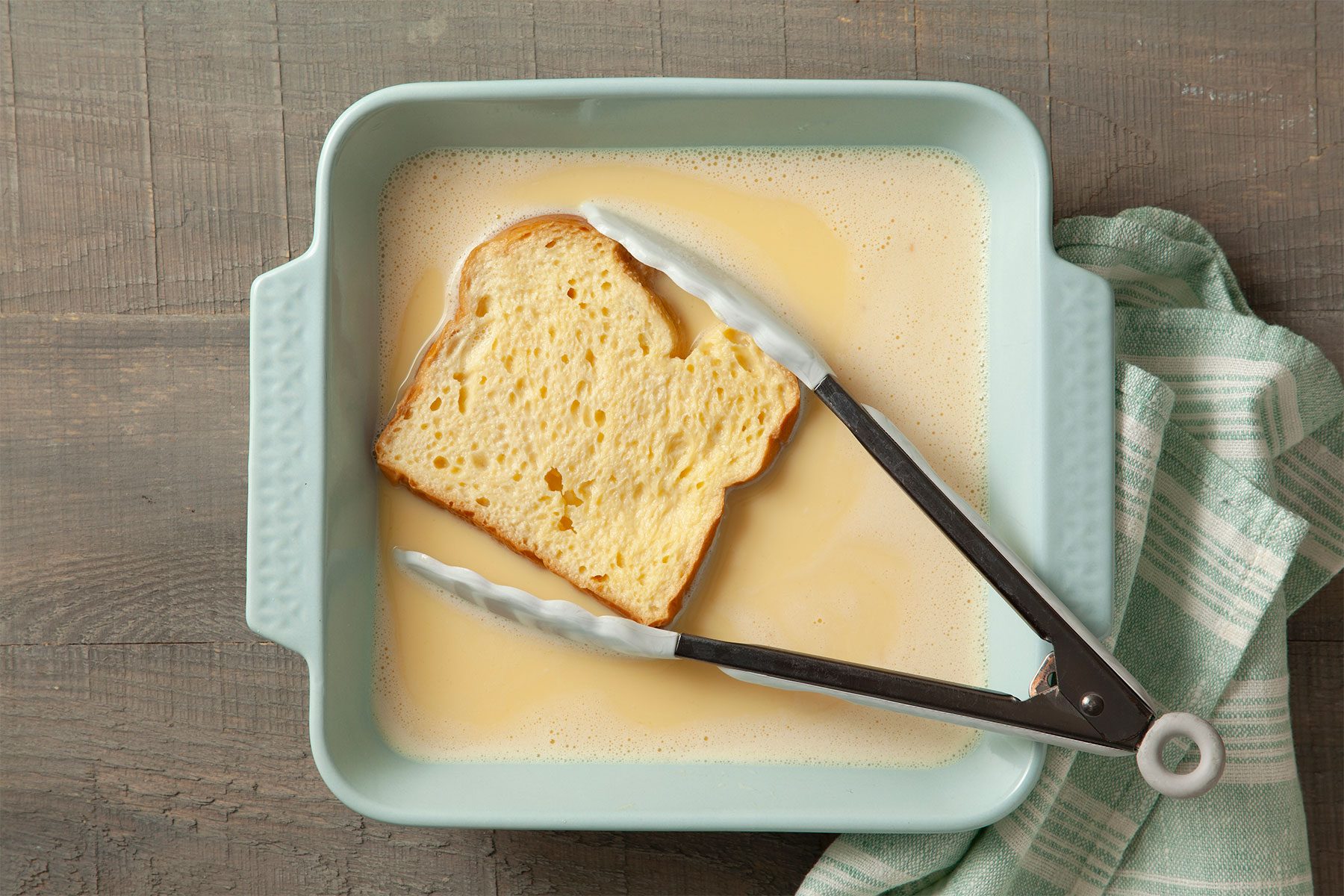 Overhead shot of dip breads one by one into egg mixture; soaked 5 seconds on each side; shallow dish; tongs; kitchen cloth; light wooden background;