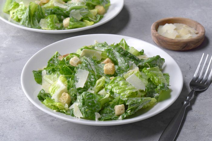 Overhead shot of Ceasar Salad; served on two white plates; sprinkle with croutons and parmesan; wooden spoon; white plate; fork; napkin; grey background