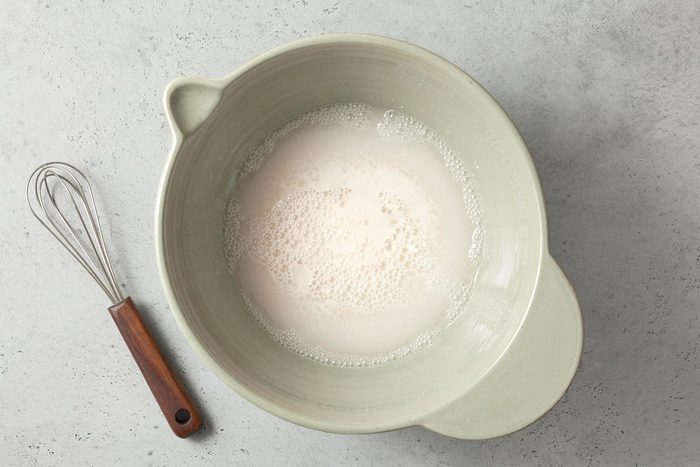 A large ceramic mixing bowl filled with a frothy liquid mixture sits on a gray countertop. Beside the bowl on the left side is a whisk with a stainless steel head and a brown wooden handle.