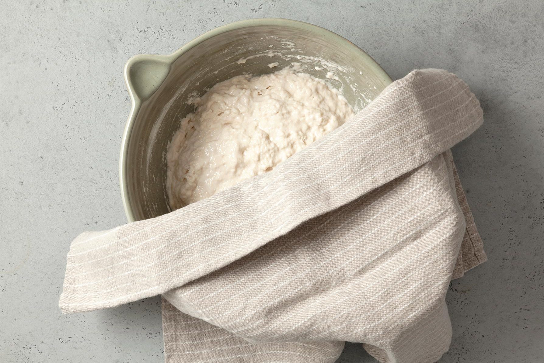 A green ceramic bowl containing dough is partially covered with a beige-striped kitchen towel. 