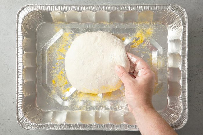 A hand holding a circular piece of pizza dough over a silver baking tray lightly dusted with flour or cornmeal.