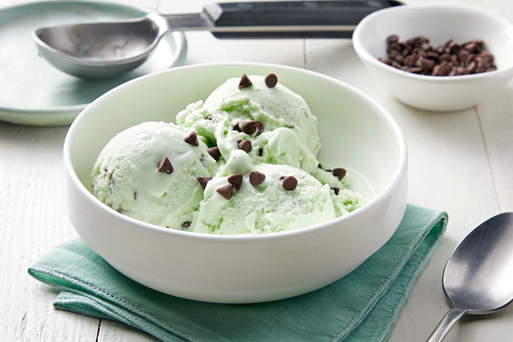 3/4th shot; white background; Mint Chocolate Chip Ice Cream served in a small bowl over kitchen cloth with silver spoon, chocolate chips in small bowl with ice cream scooper;