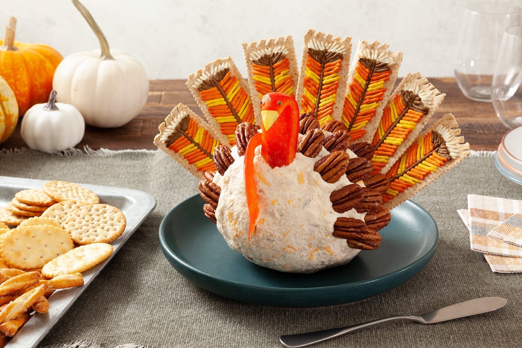 A festive cheese ball appetizer shaped like a turkey with pecans adorning the body, crackers as tail feathers, and a red bell pepper slice for the wattle, displayed on a dark green plate. In the background, there are crackers on a separate plate and a white pumpkin.