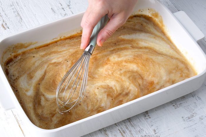 A hand using a whisk to stir a creamy, light brown mixture in a white rectangular baking dish placed on a distressed white wooden surface.