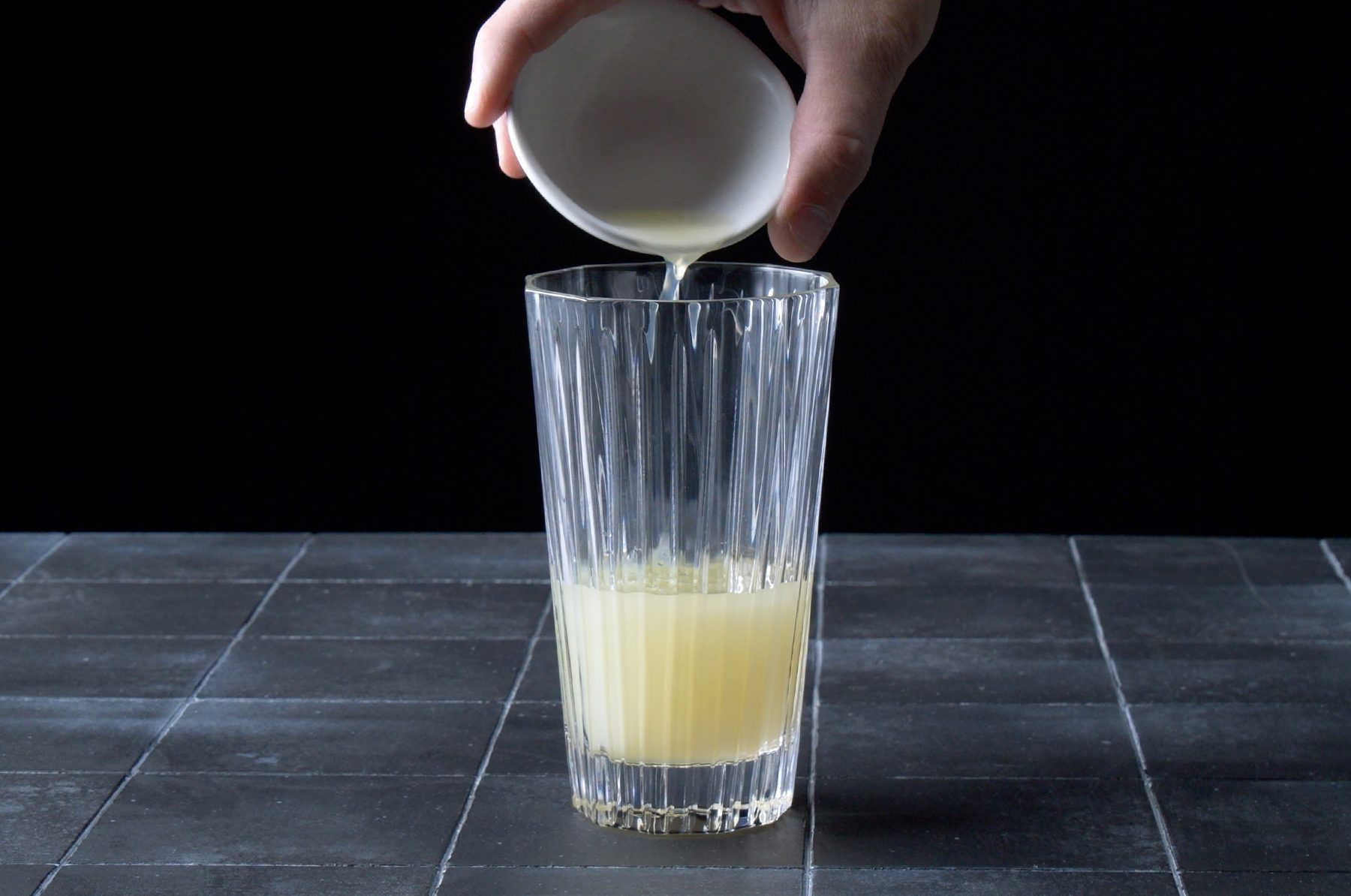 A hand is pouring a light yellow liquid from a small white cup into a tall, clear glass that is partially filled with the same liquid.