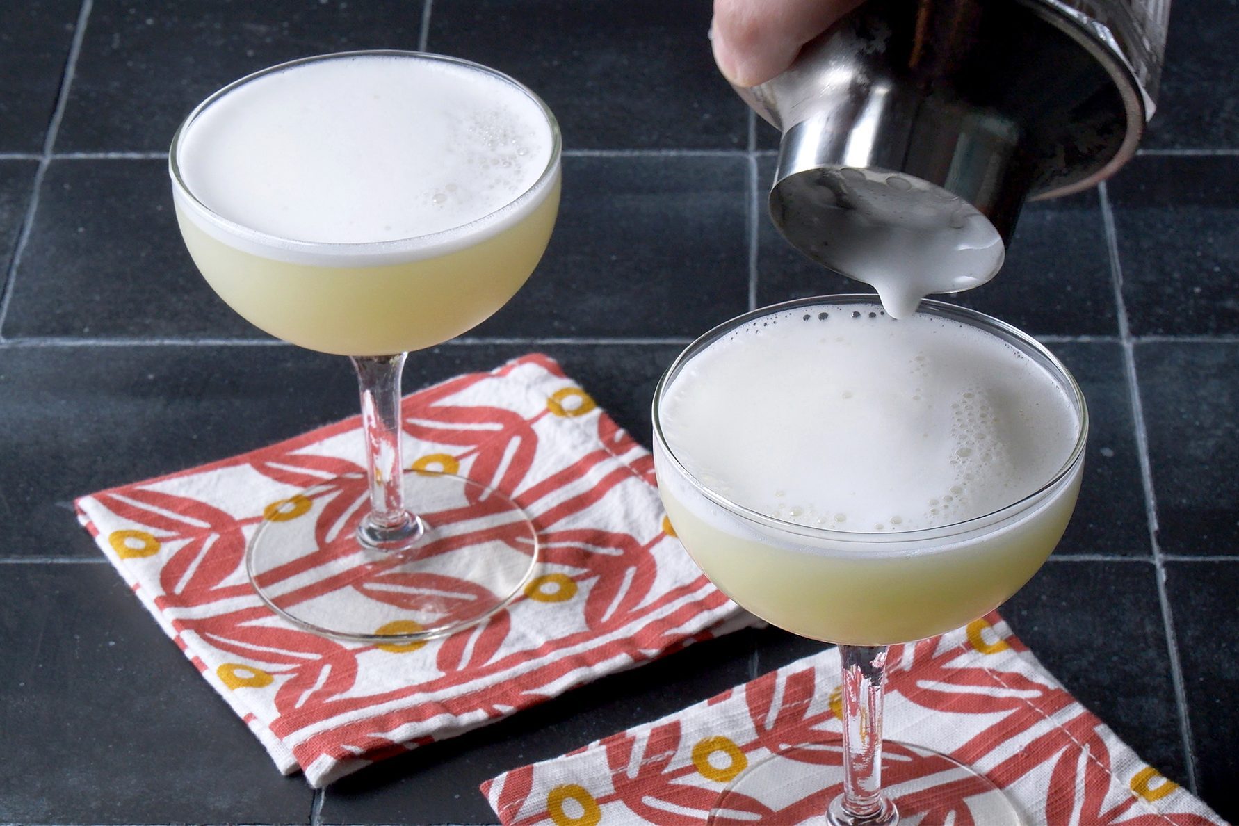 A person pours a foamy white cocktail from a shaker into one of two coupe glasses on a black tiled surface.