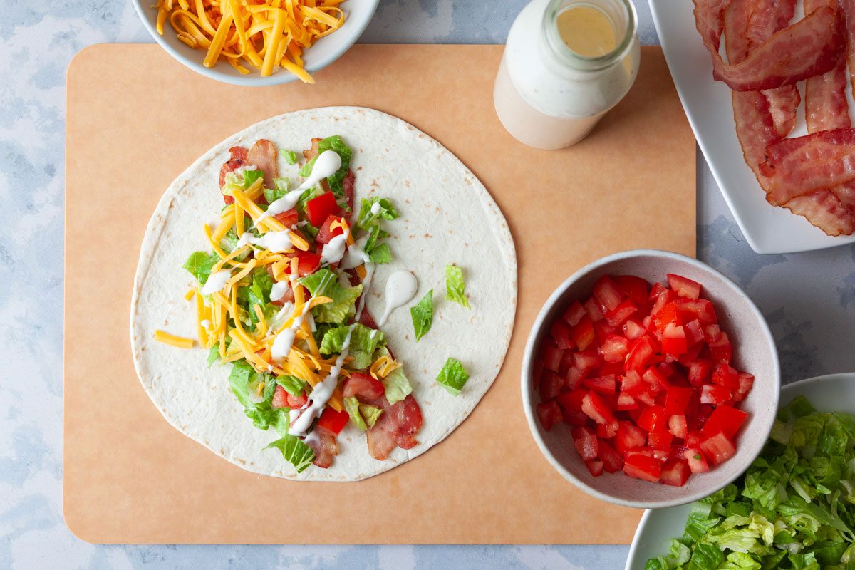 Assembly station for Taste of Home BLT Wraps, one wrap being in the process of being built by layering bacon, lettuce, tomato, cheese and ranch dressing