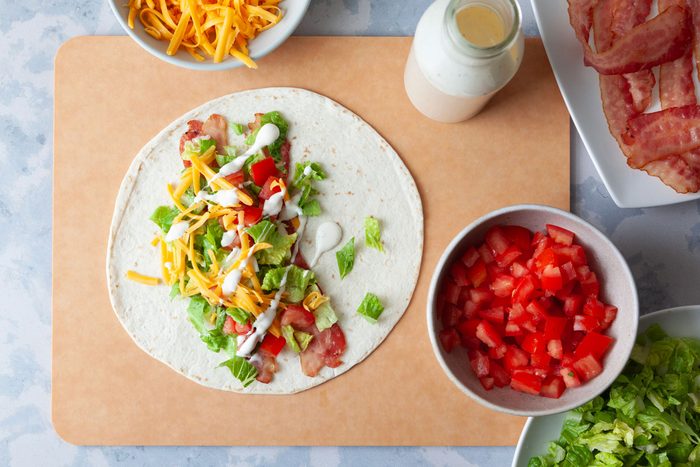 Assembly station for Taste of Home BLT Wraps, one wrap being in the process of being built by layering bacon, lettuce, tomato, cheese and ranch dressing