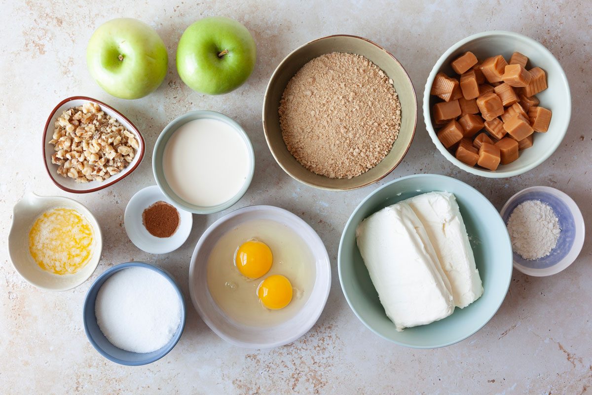 Ingredient shot of Taste of Home Caramel Apple Cheesecake including green apples, cream cheese, pecans, caramels, evaporated milk, cinnamon graham cracker crumbs, spirngform pan, eggs, cinnamon, flour, sugar, butter