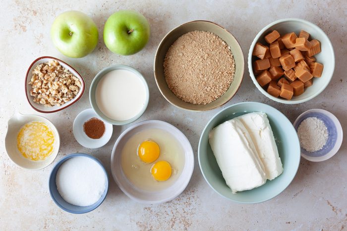 Ingredient shot of Taste of Home Caramel Apple Cheesecake including green apples, cream cheese, pecans, caramels, evaporated milk, cinnamon graham cracker crumbs, spirngform pan, eggs, cinnamon, flour, sugar, butter