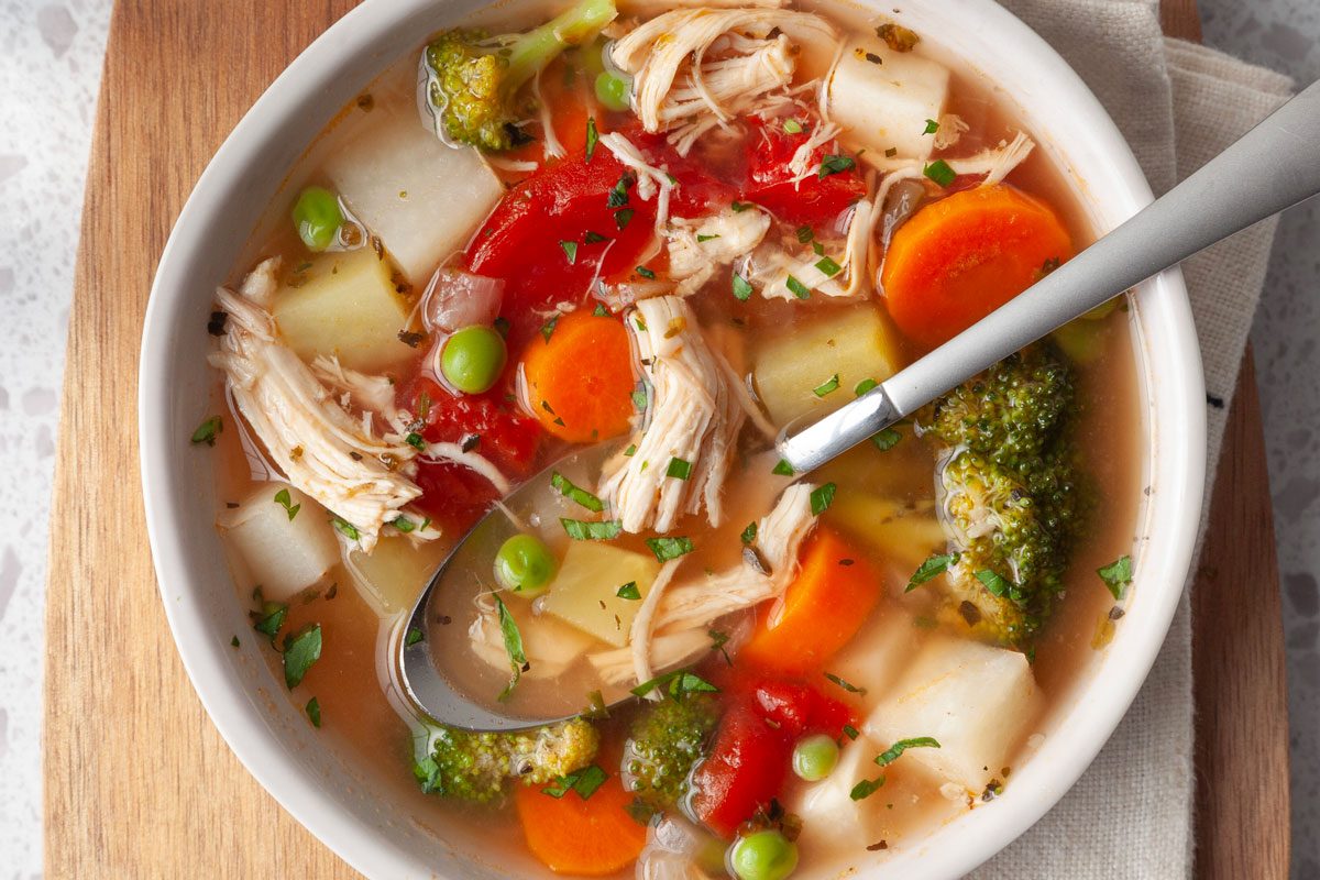 Single serving of Taste of Home Chicken Vegetable Soup in a white bowl on a wooden cutting board and a beige linen with a spoon