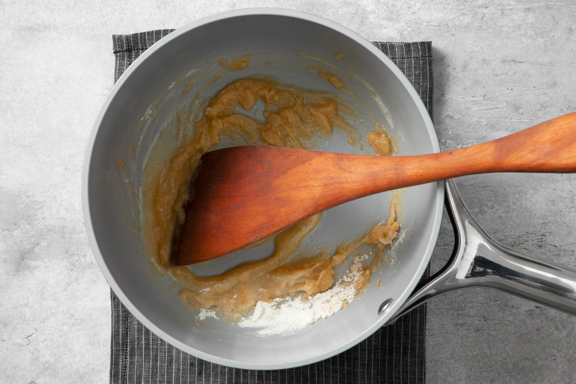 Overhead shot of a large saucepan; melt butter over medium heat; stir in flour and seasonings until smooth; wooden spatula; kitchen cloth; grey marble background;