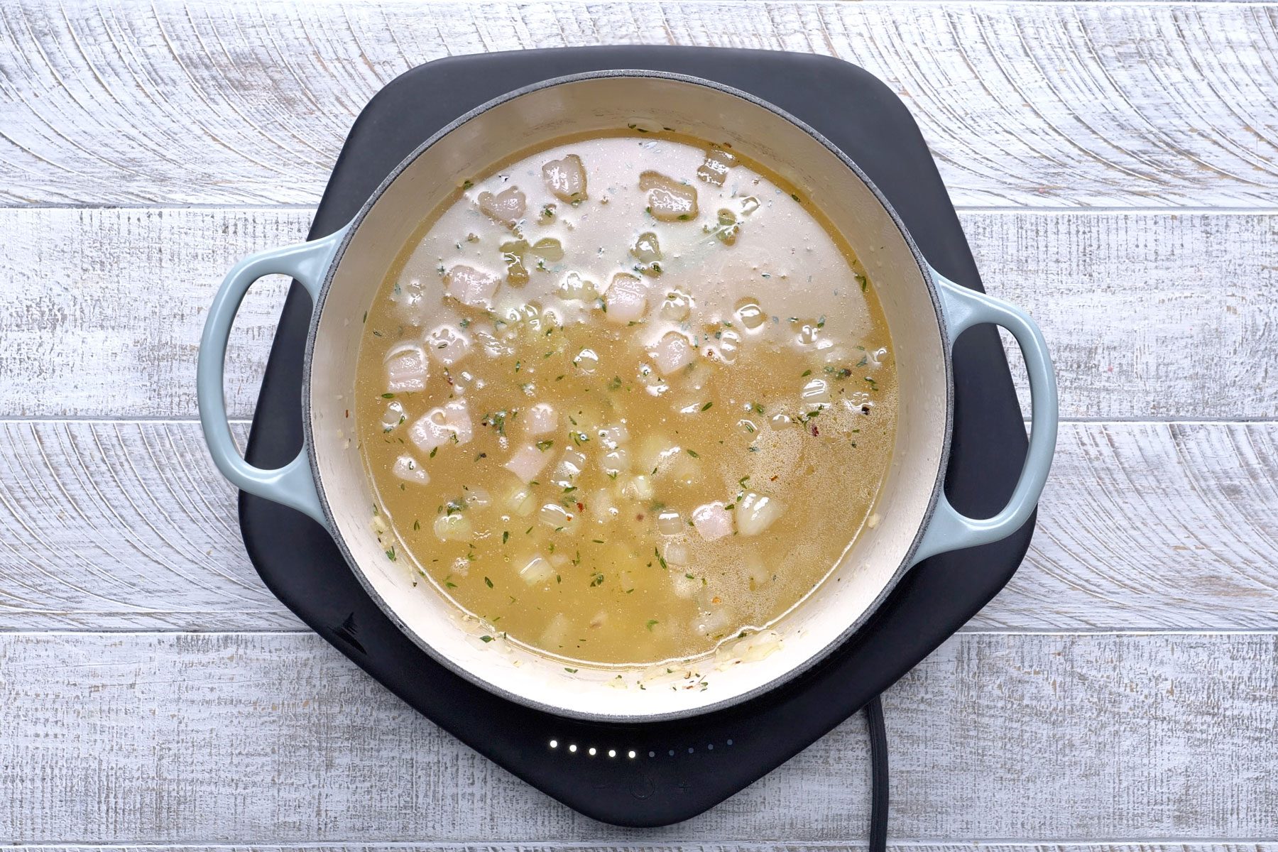 Pork belly, garlic, bay leaves, thyme, red pepper flakes and pepper simmering in a light broth in a dutch oven against a textured background.