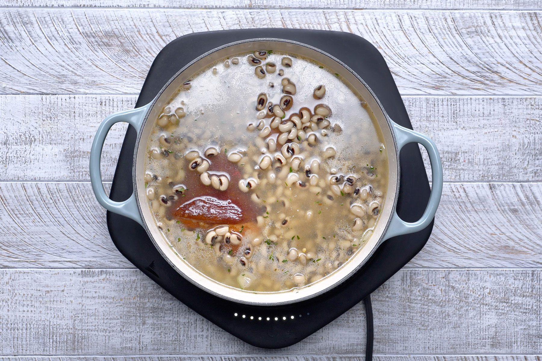 A pot of black-eyed peas simmering with ham hocks in a broth on an electric stove.