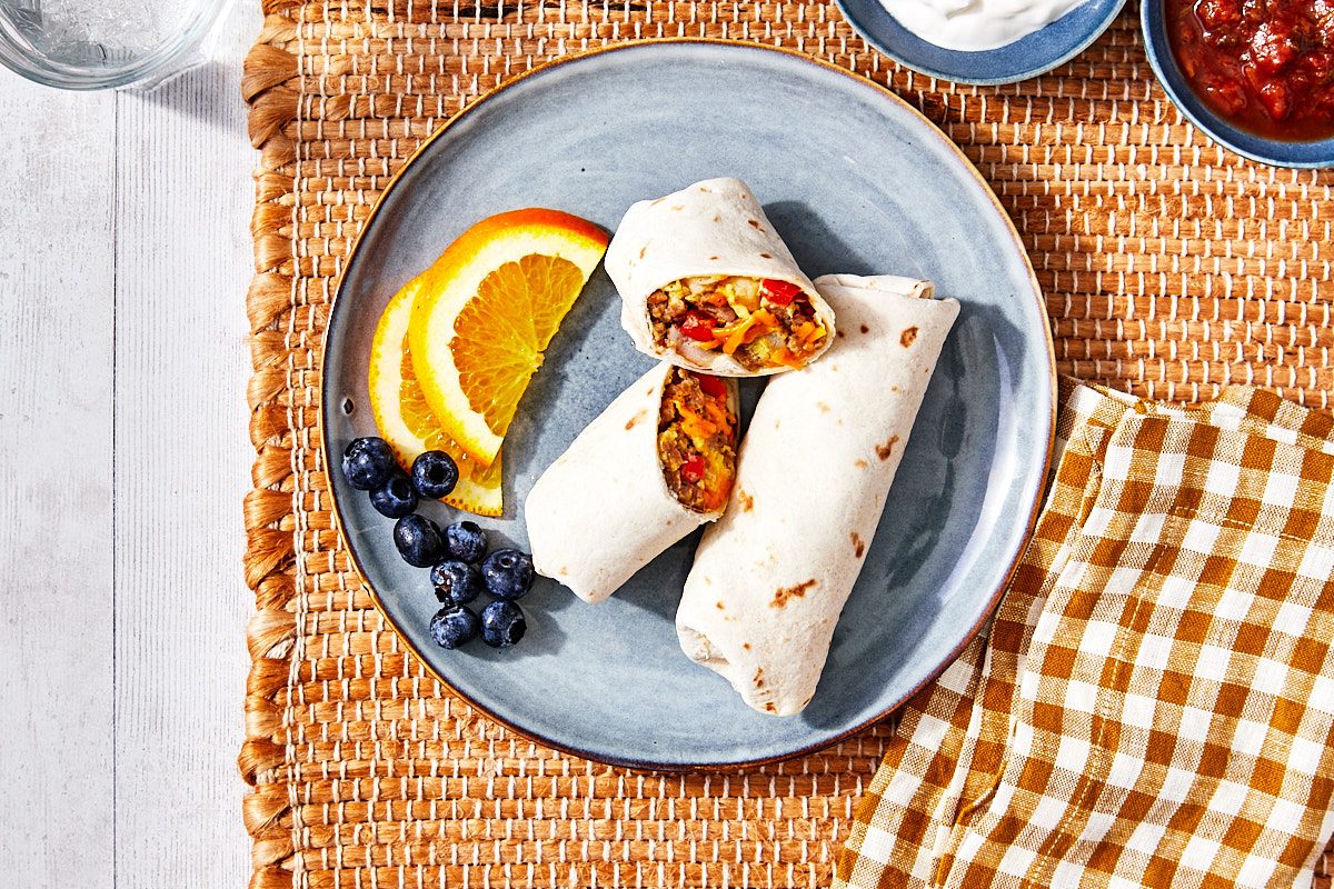 Freezer Breakfast Burritos Hero- Two burritos plated with fruit, one burrito cut in half with filling exposed.