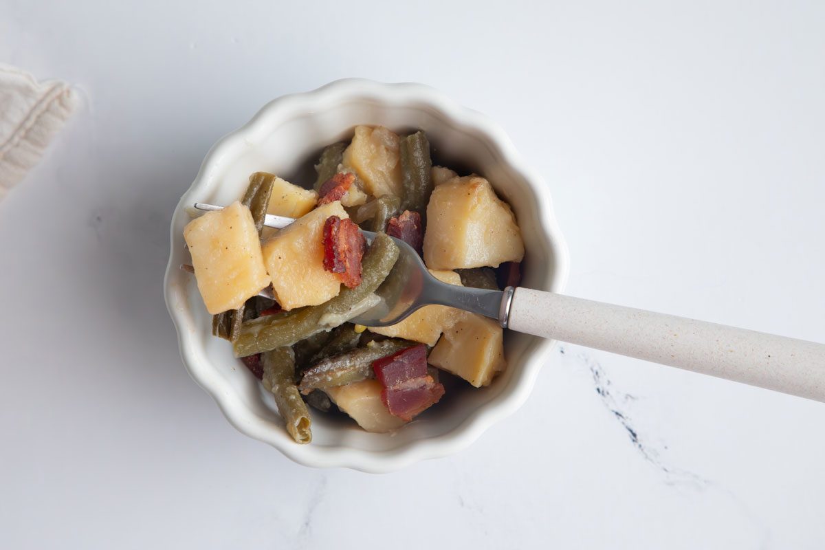 Horizontal beauty shot for Taste of home green beans and potatoes in small bowl with fork holding food on a marble surface.