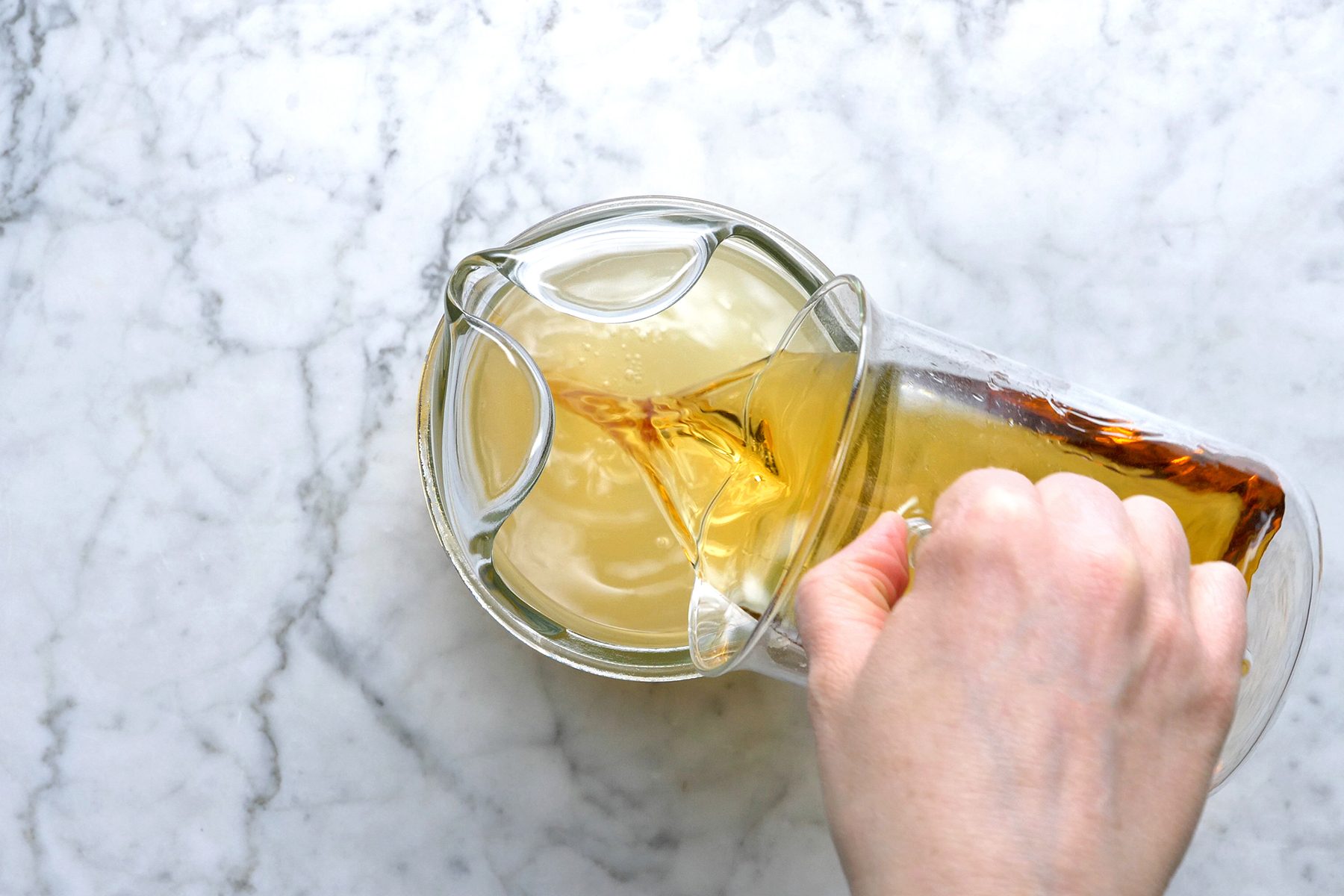 A hand is seen pouring brown liquid from a glass container into a circular glass bowl filled with a light yellowish liquid. 