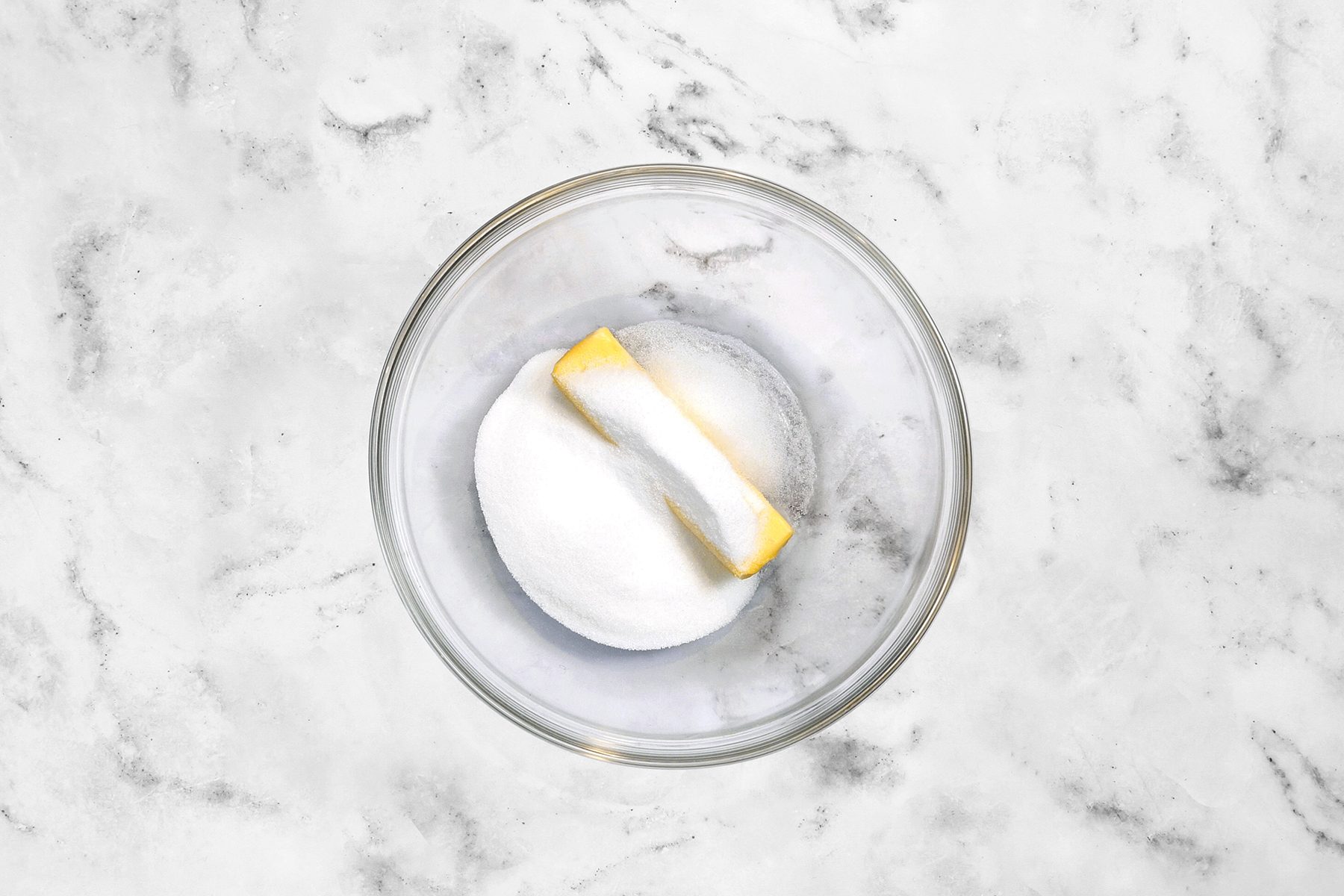 A clear glass bowl placed on a marble countertop contains an unmelted stick of butter and a mound of granulated sugar.