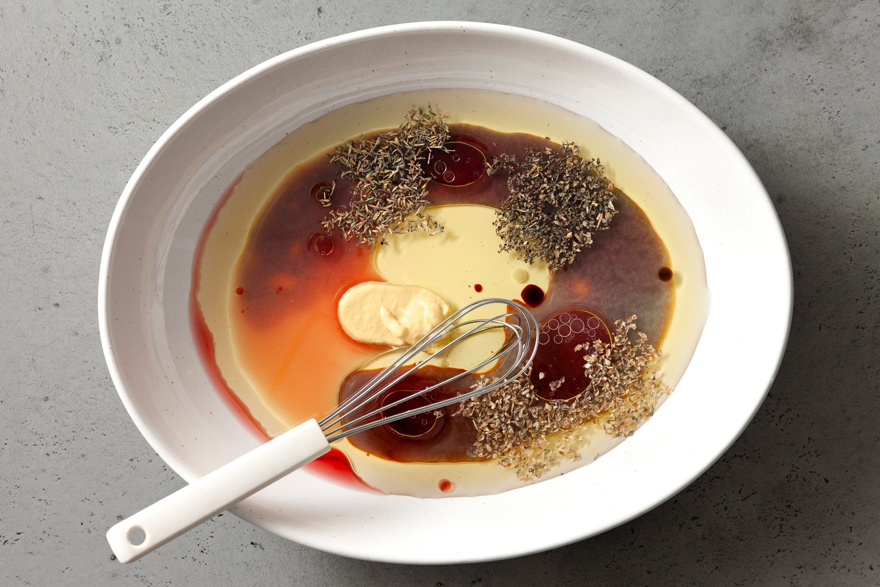 Olive oil, lemon juice, red wine vinegar, Worcestershire, soy sauce, mustard, basil, oregano and thyme are in a white bowl. A whisker rests at the edge of the bowl.