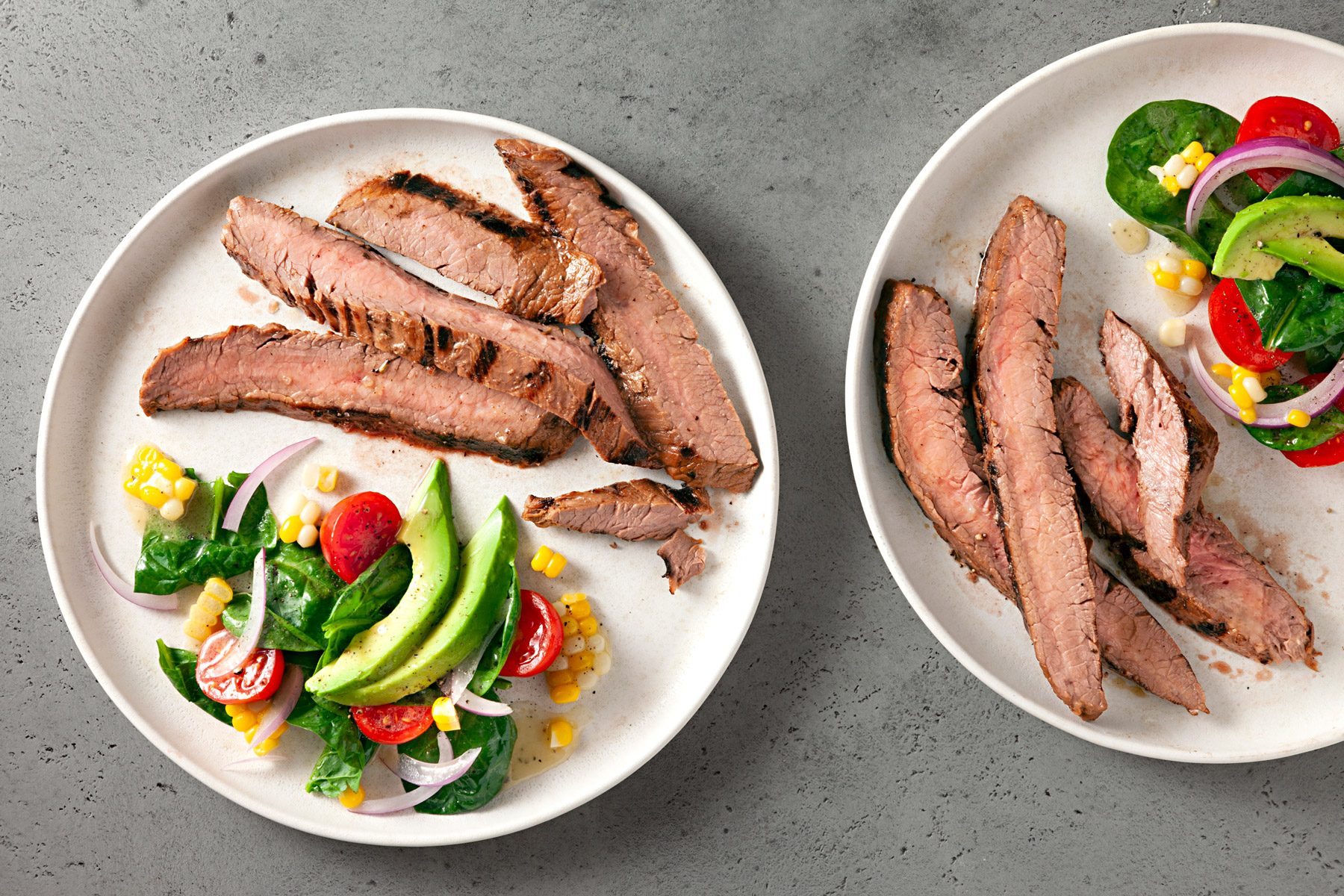 Two plates containing sliced grilled steak and mixed salad are displayed against a textured grey background.
