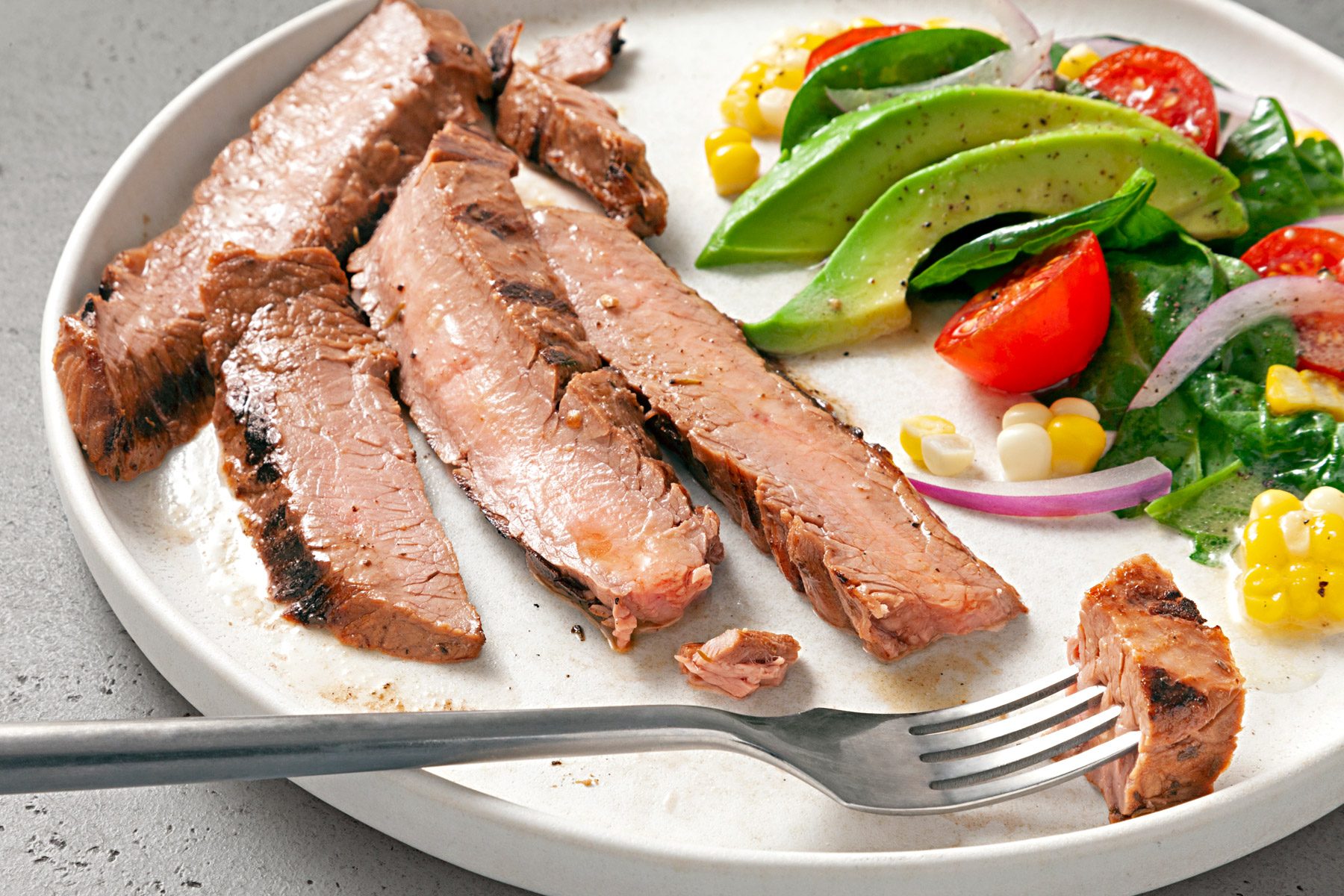 A plate of grilled beef strips served with a side of fresh vegetables, including sliced avocado, tomato halves, red onion slices, corn kernels, and spinach. A fork is picking up a piece of the beef.